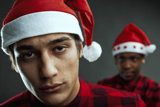 Two young men wearing festive santa hats and checkered red shirts are gazing seriously at the camera, capturing a moment of friendship and holiday spirit indoors against a gray background