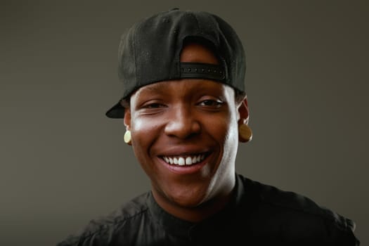 Portrait of a cheerful young black man wearing a baseball cap and ear plugs, smiling confidently at the camera against a gray background, radiating joy and style