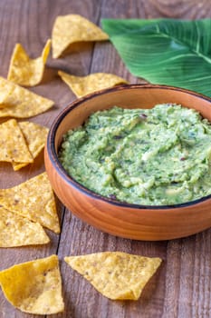Wooden bowl of guacamole with tortilla chips and a large green leaf on a rustic wooden table