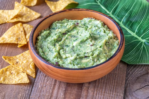 Wooden bowl of guacamole with tortilla chips and a large green leaf on a rustic wooden table