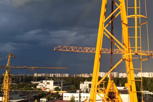 Tower cranes against the sky with clouds and rainbow.