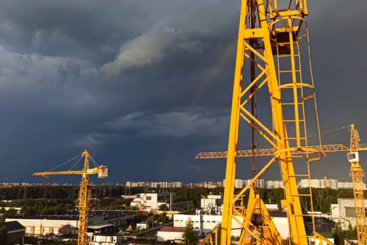 Tower cranes against the sky with clouds and rainbow.