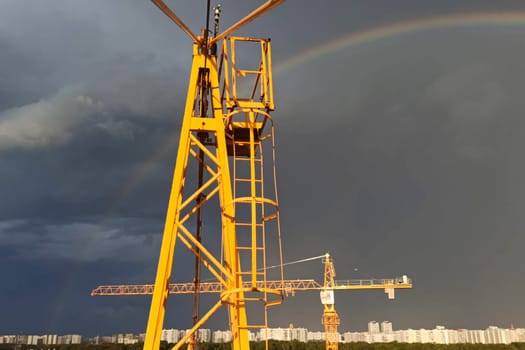 Tower cranes against the sky with clouds and rainbow.