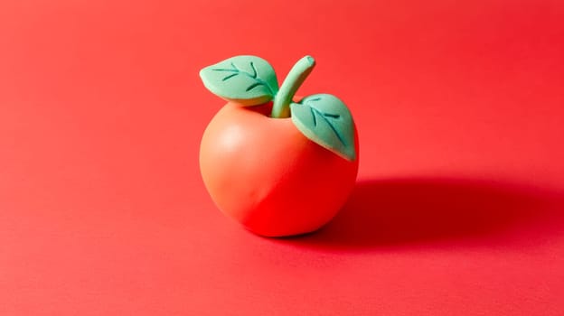 Shiny plasticine apple with green leaves and stem is casting a shadow on a vibrant red background, creating a playful and artistic still life