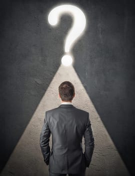 Rear view doubtful businessman inside a dark room, stands in front of a concrete wall with illuminated interrogation mark. Seeking answer to his questions. Contemplation concept