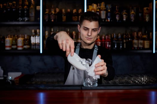 Barman puts the ice cubes into a glass with alcoholic drink at the bar