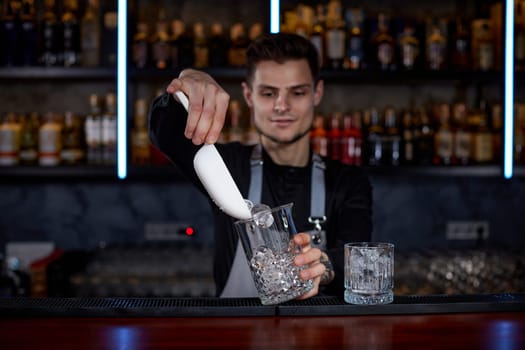 Barman puts the ice cubes into a glass with alcoholic drink at the bar