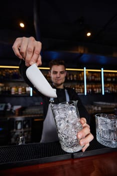 Barman puts the ice cubes into a glass with alcoholic drink at the bar