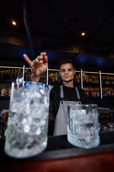 Barman in apron stirring ice cubes in cocktail glass with spoon at the bar
