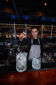 Barman in apron stirring ice cubes in cocktail glass with spoon at the bar