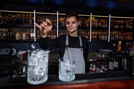 Barman in apron stirring ice cubes in cocktail glass with spoon at the bar