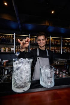 Barman in apron stirring ice cubes in cocktail glass with spoon at the bar