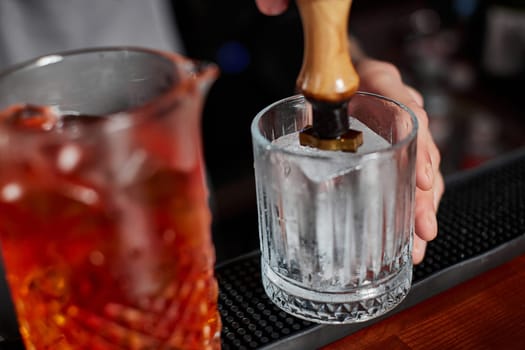 barman in apron pours old fashioned cocktail into glass with large piece of ice on the bar counter.