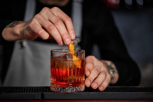 bartender makes a cocktail in a glass with orange peel at the bar, close-up