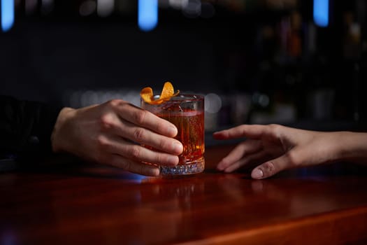 Barman holding out glass with alcoholic cocktail to the client. Bartender is making cocktail