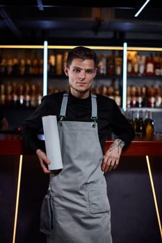 Handsome barman in apron looking at camera and holding box for bottle at the bar counter