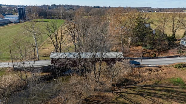 A covered bridge stands along a minor road, surrounded by bare trees and grassy fields. The serene rural landscape shows hints of early spring as foliage starts to awaken.