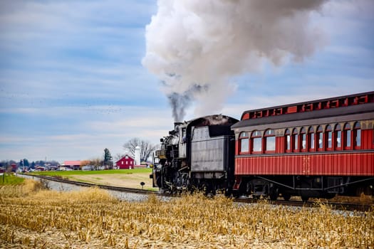A steam train chugs along a country track amidst golden fields, releasing billowing smoke into the blue sky. Nearby, a classic red passenger car follows closely.