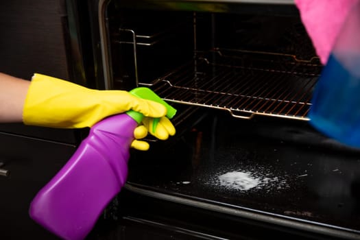 A person is diligently cleaning the oven with a purple spray bottle while wearing bright yellow gloves for protection