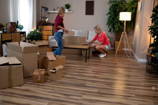 Two friends are happily unpacking boxes and carefully organizing furniture in a cozy living room space together