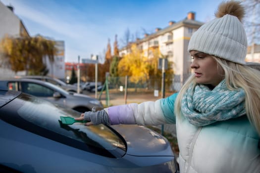 A woman is diligently cleaning the frost off her car window, showcasing the typical winter conditions in vibrant colors