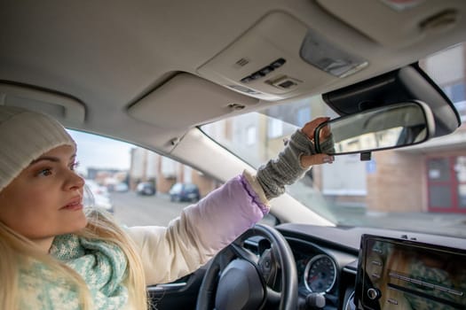 A woman wearing a cozy beanie and a stylish scarf is adjusting her cars rearview mirror while being parked