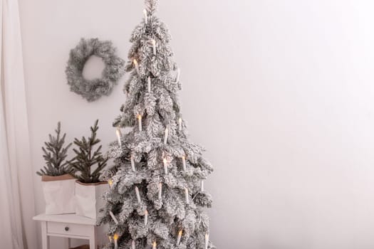 Snow-covered artificial spruce with an electric garland in the form of candles, standing against a white wall, at room. Copy space.
