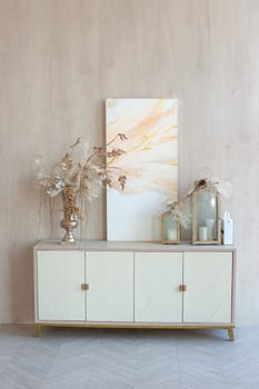 a white chest of drawers with an abstract canvas, a vase with Christmas decor and decorative lanterns, stands against a beige wall, in a neutral interior