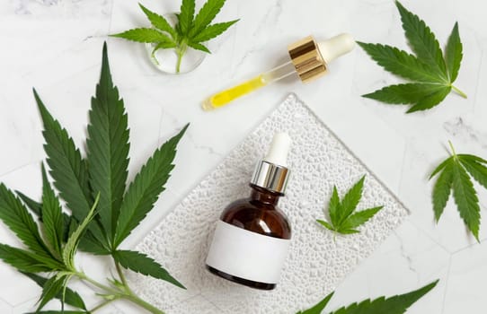 Dropper bottle with blank label near green cannabis sativa leaves on a marble table top view. Cosmetic Mockup, Copy space. Organic skincare beauty product. Eco friendly CBD oil