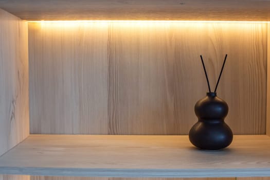 On a wooden shelf with warm lighting there is a small black ceramic vase with two sticks of incense. Copy space