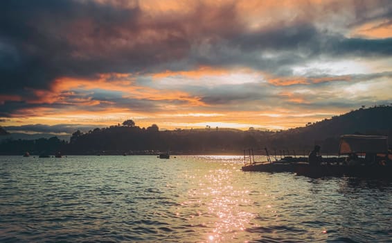 This photograph captures a beautiful sunset over Patratu Lake in Patratu Valley, India, as seen from a boat. A dock or platform is visible in the distance, and the golden light reflects off the water, creating a serene and picturesque scene. High-resolution image available for digital download. Patratu Lake Sunset from Boat, Dock View in Patratu Valley, India. High quality photo