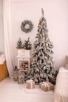 Snow-covered artificial christmas spruce with an electric garland in the form of candles, and craft gift box, standing at bedroom
