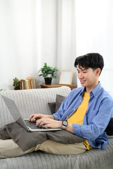 Smiling young Asian man working or browsing on a laptop in a cozy living room.