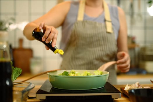 Woman preparing a healthy meal in a kitchen, adding soy sauce into a pan.