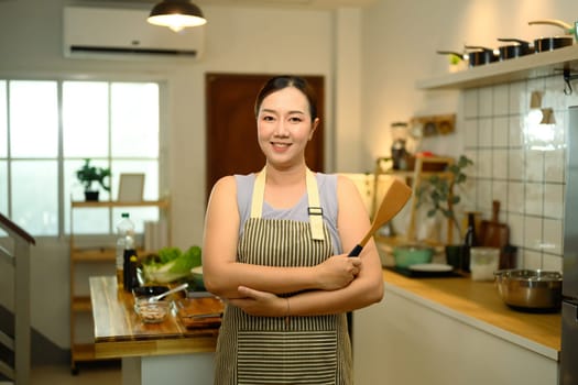 Portrait of smiling adult woman posing with a wooden spoon in her kitchen, ready to cook.