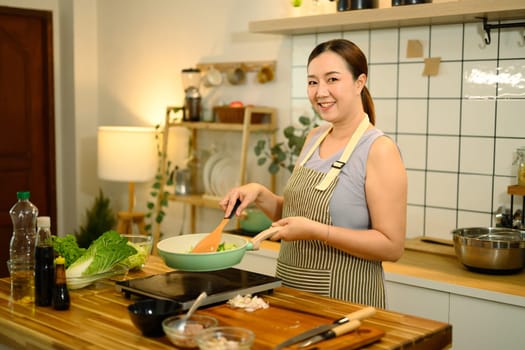 Smiling 40s asian woman wearing apron preparing a healthy meal in a modern kitchen.