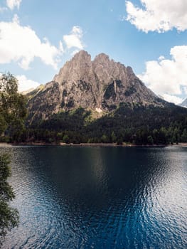 spectacular landscape of calm lake surrounded by massive green mountains and coniferous trees against blue sky in Aiguestortes i Estany of Saint Maurici National Park