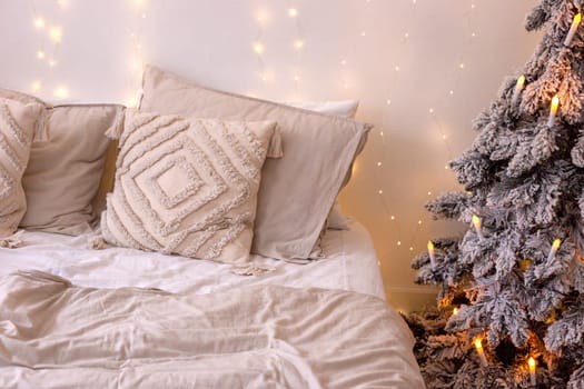 A bed with pillows and a beige bedspread, next to a snow-covered artificial Christmas tree with an electric garland in the form of candles, standing in the bedroom