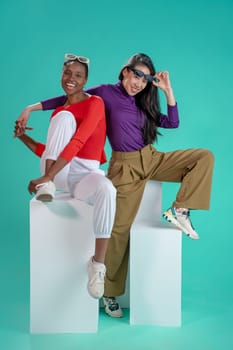Two stylish women pose playfully in a colorful studio, showcasing the latest fashion trends with flair