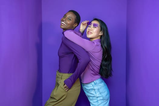Two models pose playfully against a vibrant purple background, showcasing bold fashion and expressions.