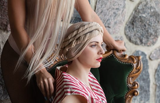 A stylist carefully prepares a unique hairstyle on a seated model. The rich textures of the stone wall complement the vintage chair, adding elegance to the creative process in soft lighting.