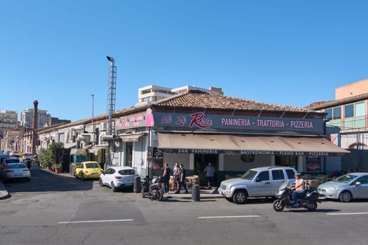 Catania. Italy - February 20, 2025: Street view of a marketplace in Catania, Sicily, featuring local businesses such as a fruit store, a car workshop, and a reflexology clinic on a sunny day.
