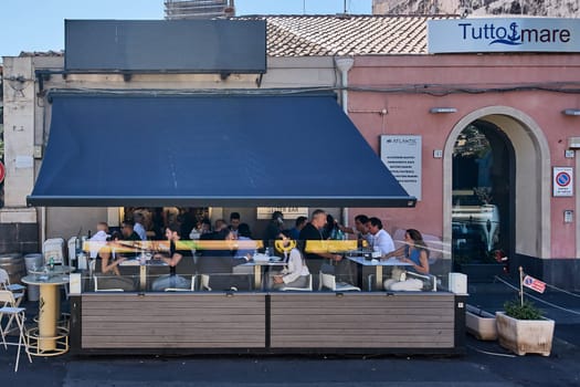 Catania, Italy - February 20, 2025: Lively dining scene at Tutto Mare restaurant with outdoor seating, glass panels, and diners enjoying their meals. Pink facade and potted plants visible. Concept: urban dining