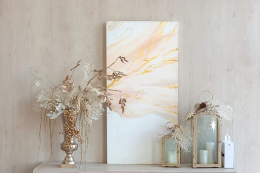 an abstract canvas, a vase with Christmas decor and decorative lanterns with candles stand on a table against the wall. Close up