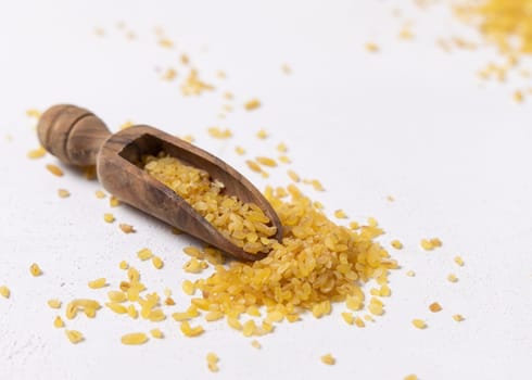 Raw dry uncooked yellow bulgur grain in a wooden scoop on white table closeup. Middle Eastern cousine. Organic super food for healthy diet. Ancient alternative cereal. seeds