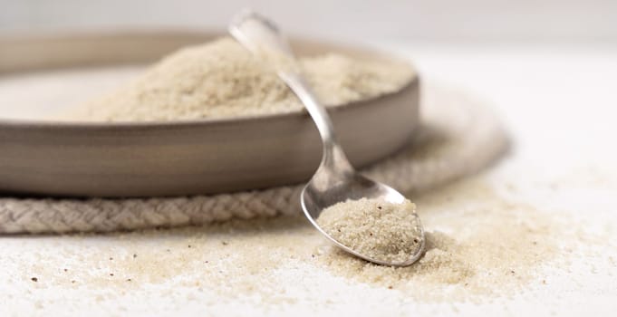 Raw uncooked fonio seeds in a plate with a metal spoon on white table close up, copy space. African ancient pseudocereal. Organic super food. Healthy food concept. Gluten free alternative seeds