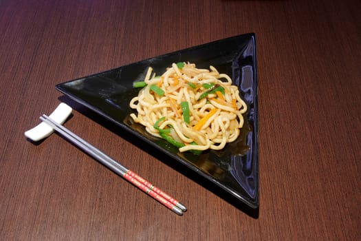 A plate of noodles with vegetables and a pair of chopsticks. The plate is triangular in shape and the vegetables are arranged in a way that makes the dish look appetizing