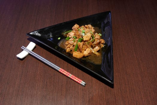 A plate of food with chopsticks on a wooden table. The plate is triangular and filled with chicken and vegetables