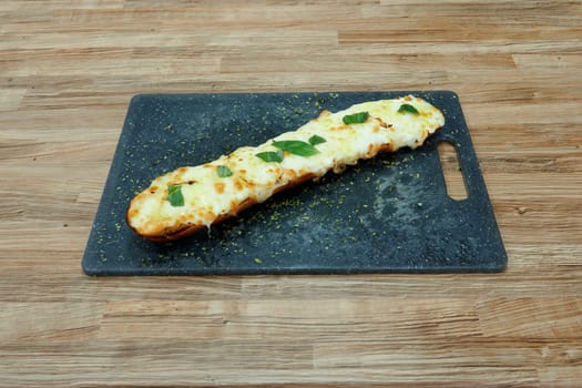 A long piece of bread with cheese and herbs on top. The bread is on a wooden cutting board