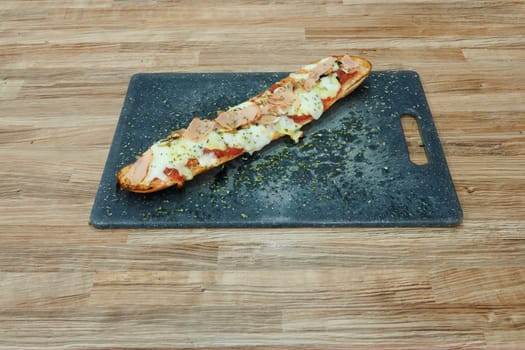 A long, thin pizza with cheese and meat toppings sits on a wooden cutting board. The pizza is cut into slices and is ready to be eaten